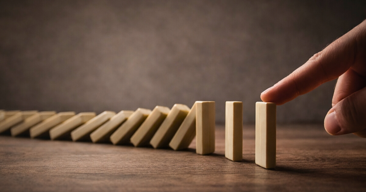 A hand pushing the first domino in a row of wooden dominoes on a table.