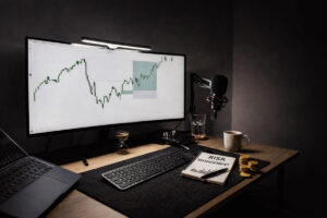 Professional stock trading setup with financial charts on screen and a notebook labeled "Risk Management" on a clean desk.