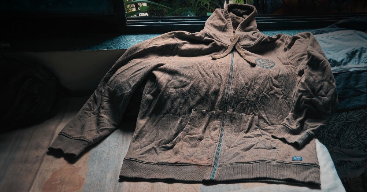 A worn olive-green oversized hoodie lying on a bed beside a window, with sunlight falling across its wrinkled fabric.